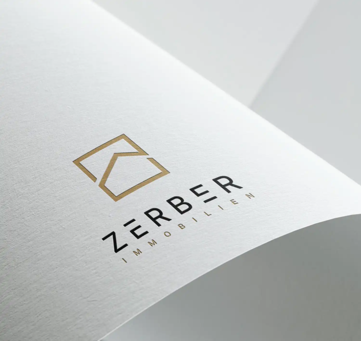 Logo der Firma Zerber Immobilien auf einem strukturierten weißen Papier. Das Logo zeigt ein minimalistisches, stilisiertes Haus in Gold mit klaren geometrischen Linien. Darunter steht der Firmenname "Zerber Immobilien" in modernem, klaren Schriftstil, wobei "Zerber" hervorgehoben ist.